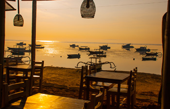 Seaside dining area with wooden chairs facing anchored boats at dusk